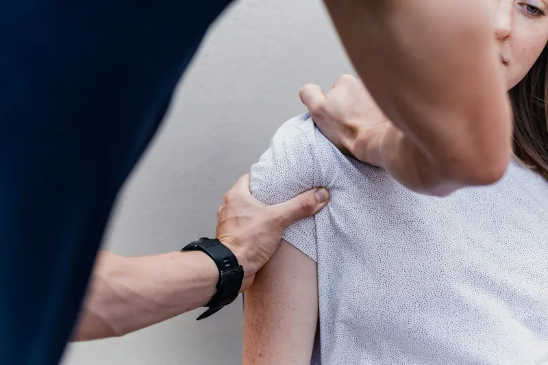 Un fisioterapeuta explora el hombro de una paciente de pie, sujetándole el brazo